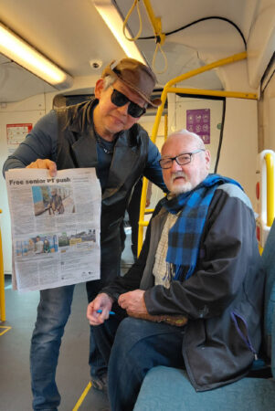 Two people are in a train carraige. One man is standing with hat and sunglasses holding up a newspaper, the other man is sitting.